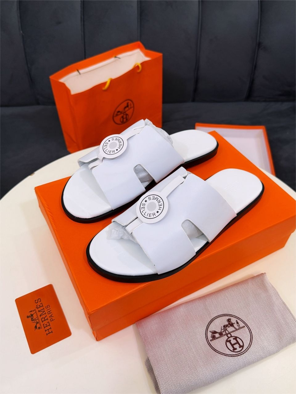 White Hermes sandals with logo placed on orange box with bag and accessories.
