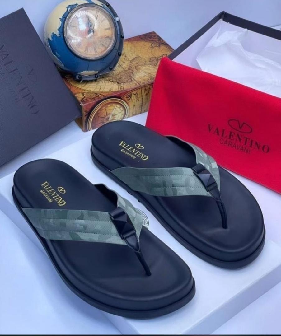 Black Valentino flip flops with camo strap and gold branding next to box and accessories.