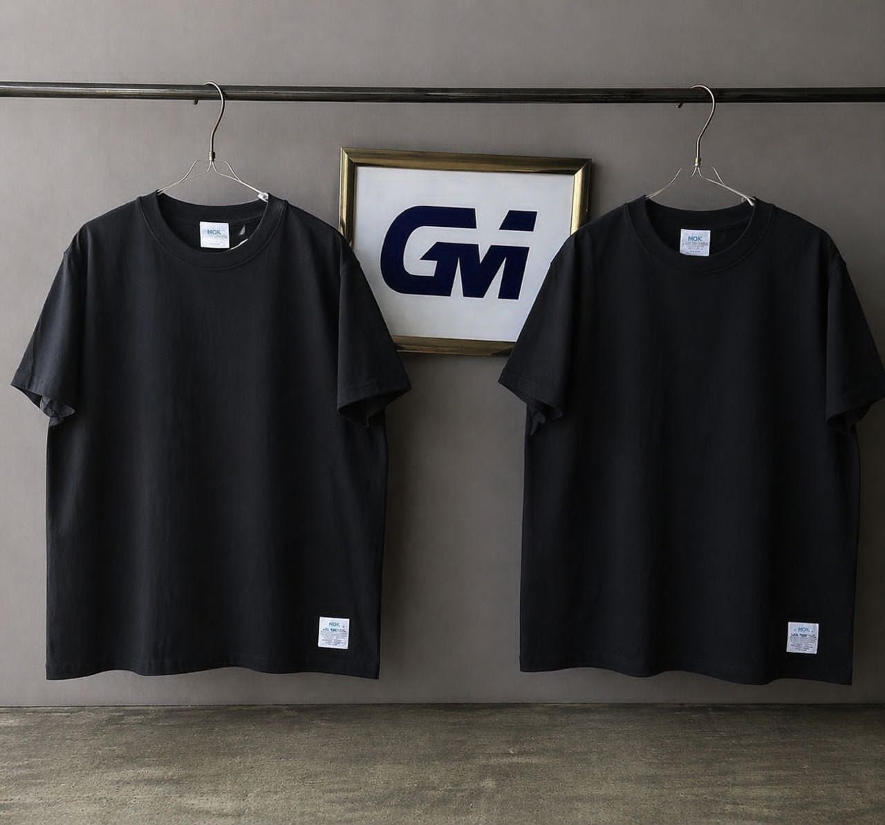 Black GM logo t-shirt displayed on hanger with framed GM sign in background.