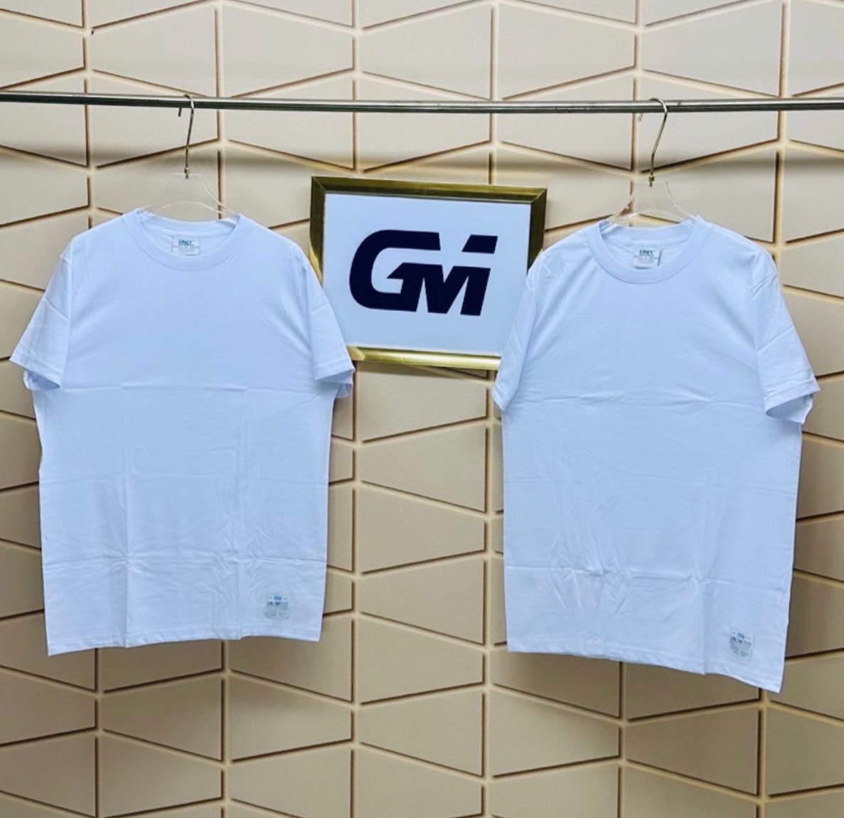 Two white plain oversized t-shirts hanging on clear hangers against a tiled wall.