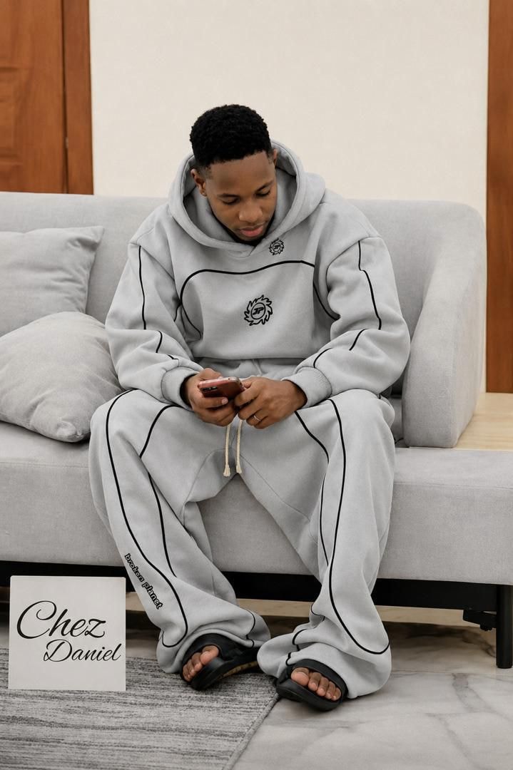 Man seated on couch wearing gray piped logo hoodie and matching pants set.