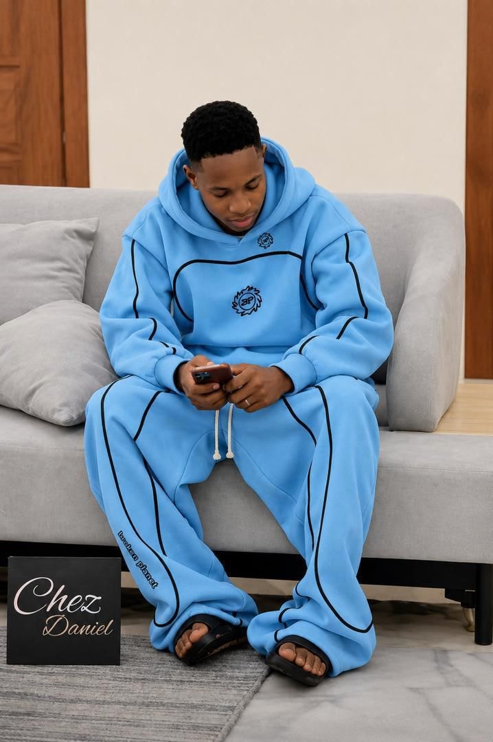 Man seated on couch wearing light blue piped logo hoodie and matching pants set.