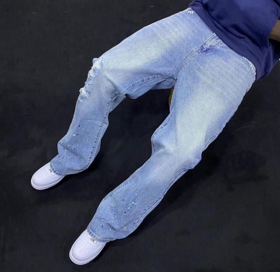 Person wearing light blue baggy jeans with knee rips, white paint splatters and white sneakers against black background.