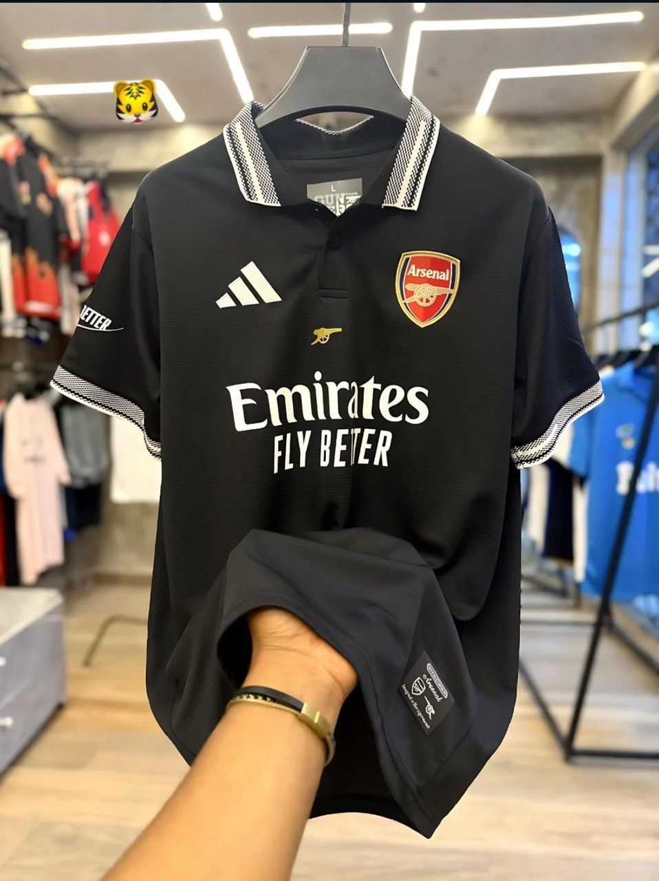 Black Arsenal Adidas Emirates football jersey held by hand in store.