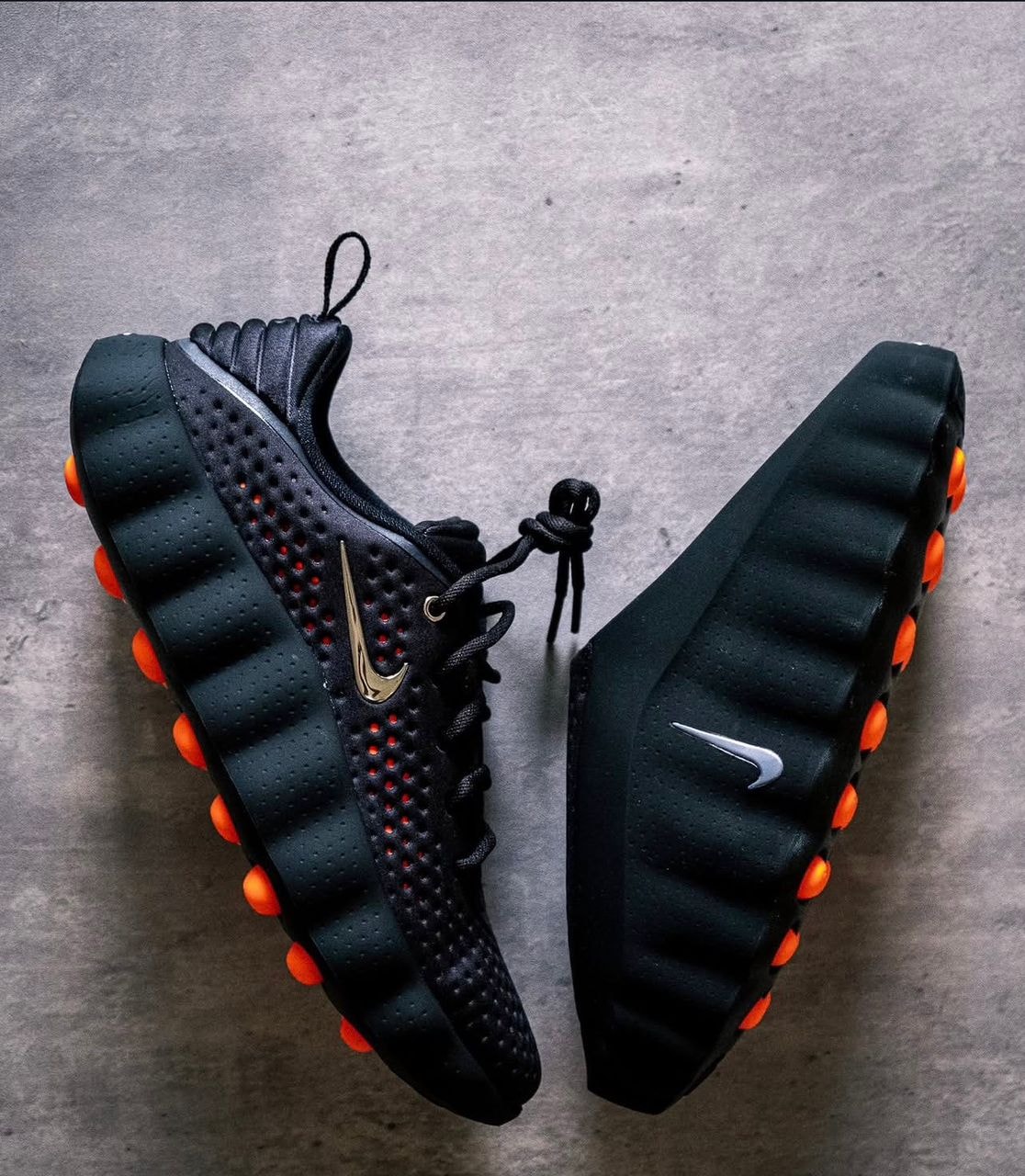 Black Nike sneakers with gold Swoosh, orange pods and textured sole on gray surface.