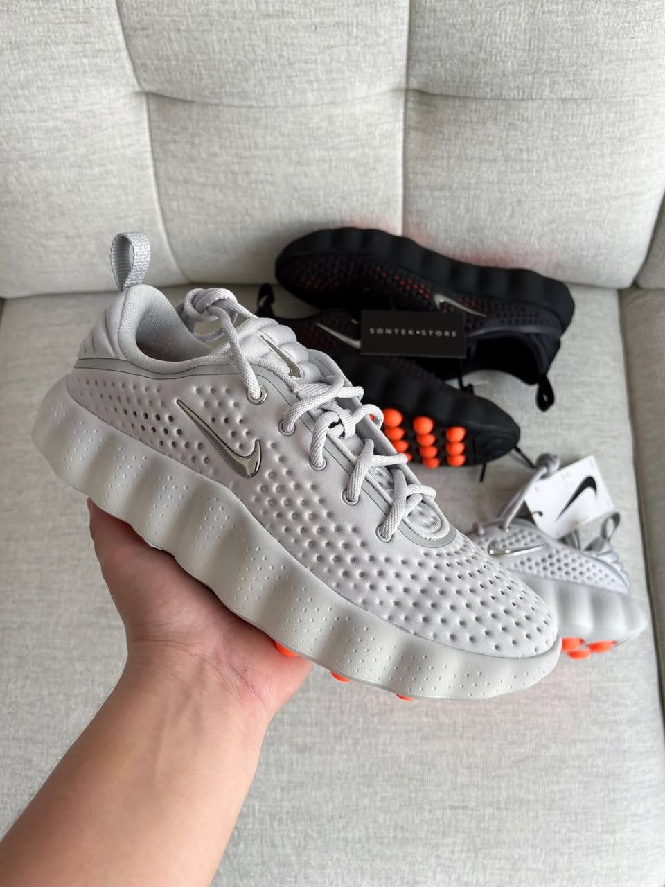 Gray Nike React sneakers held in hand showing perforated upper, orange pods and silver Swoosh.