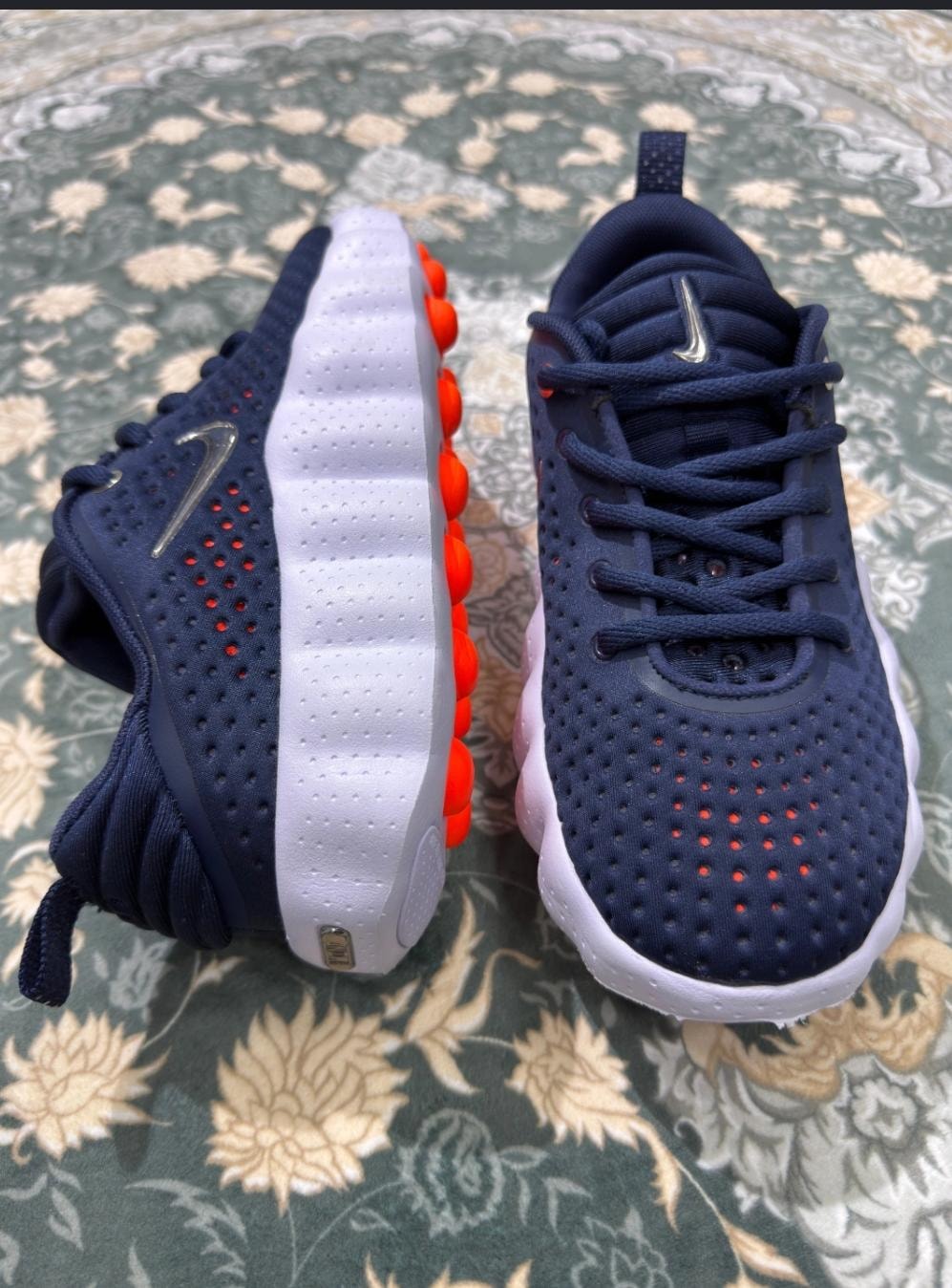 Navy blue Nike sneakers with orange sole pods and silver Swoosh on patterned rug.
