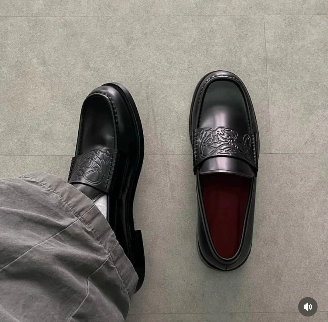 Black leather loafers with embossed design worn with grey trousers on a tiled floor.