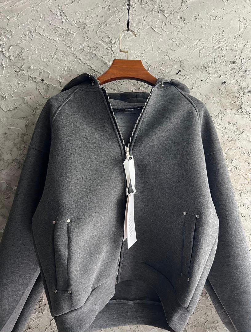 Gray zip-up hoodie hanging on wooden hanger against textured wall.