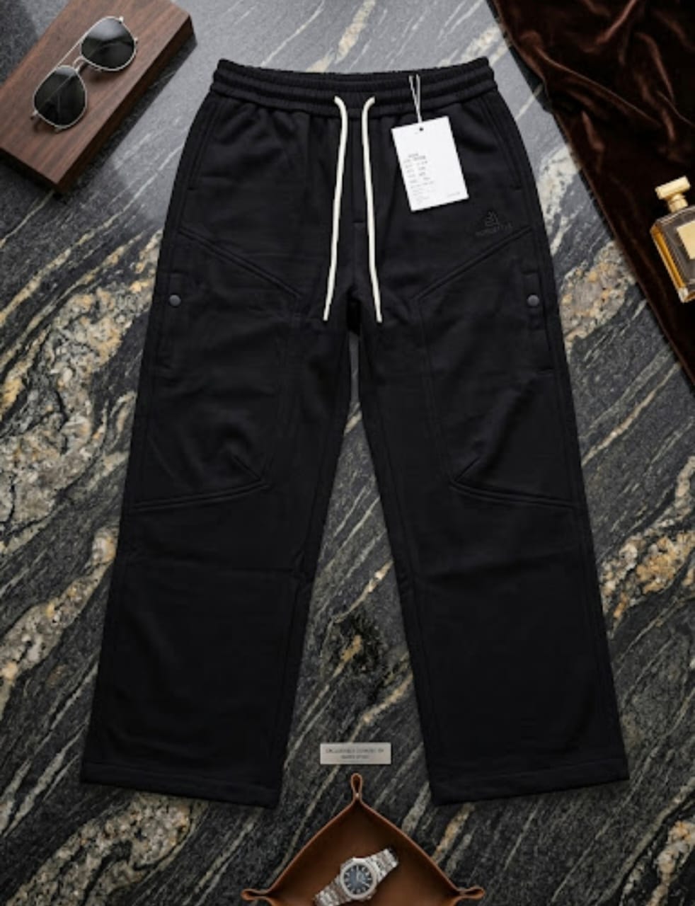 Black cargo joggers with flap pockets and tag displayed on dark marble with accessories.