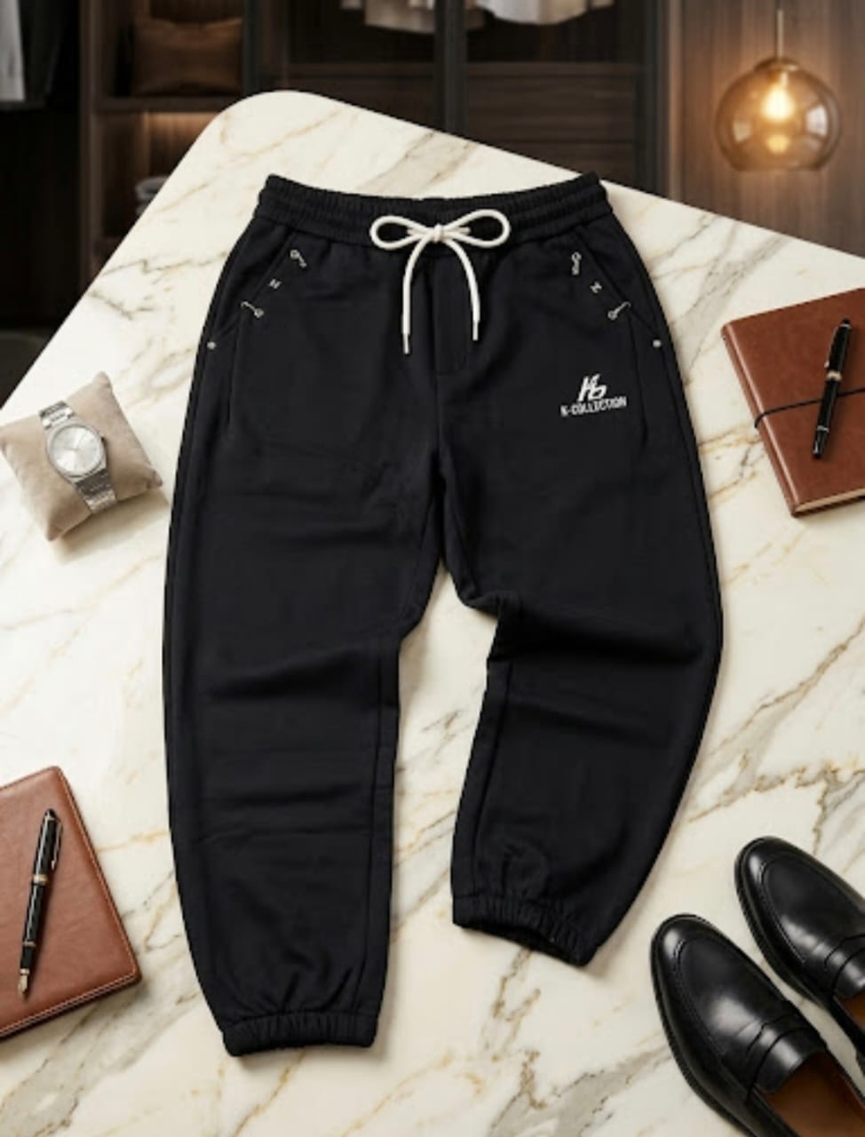 Black joggers with K-COLLECTION logo and white drawstring surrounded by notebook and shoes.