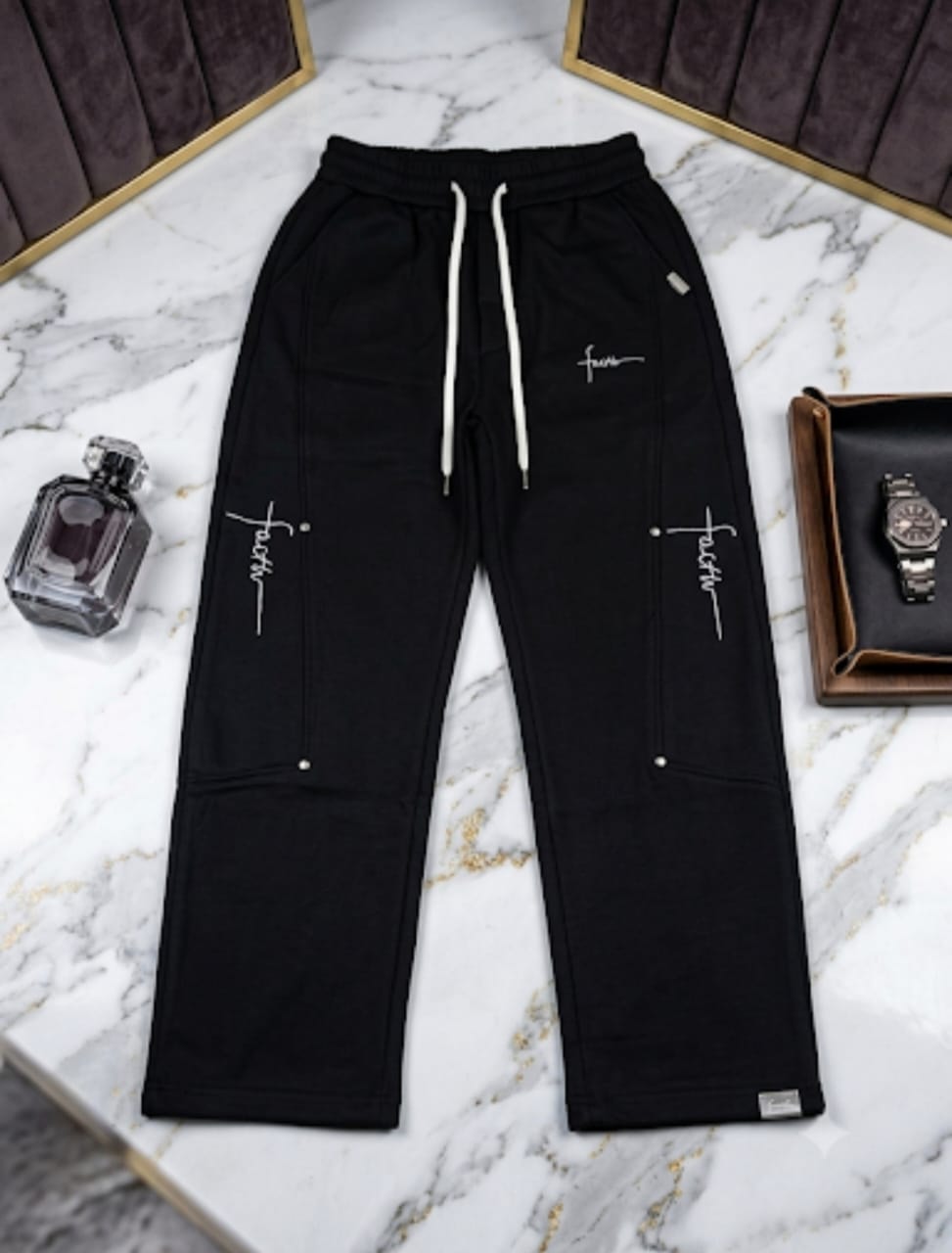 Black joggers with white faith script and cross designs on marble with perfume and watch.