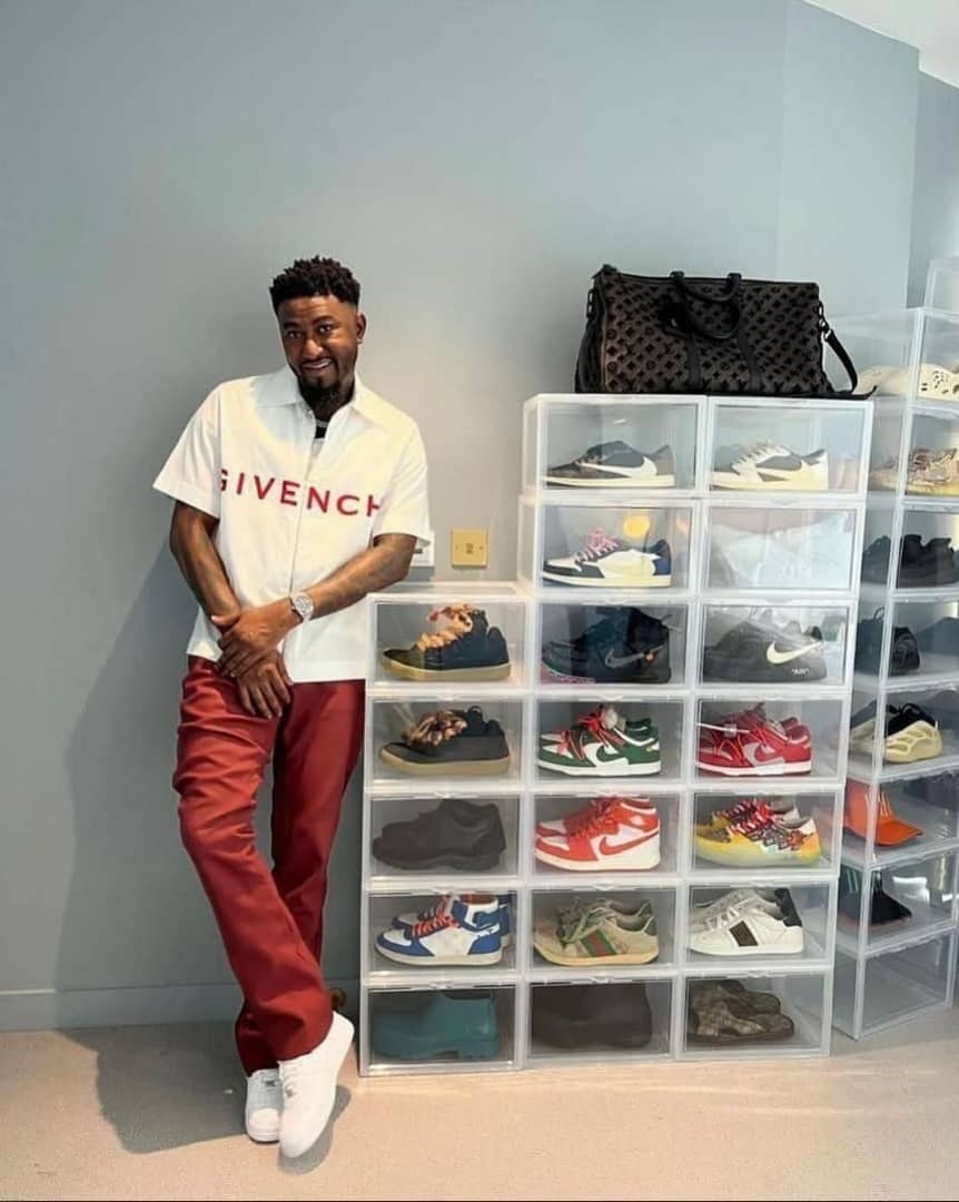 Man posing beside stacked transparent acrylic shoe boxes filled with various sneakers and a designer bag on top.