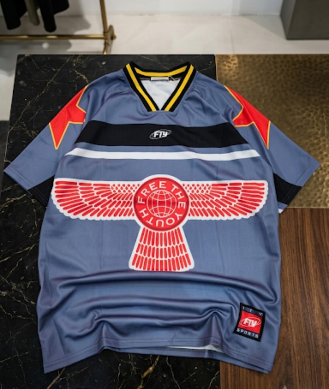 Blue graphic big tee with large red wings and Free The Youth print shown on dark marble surface.