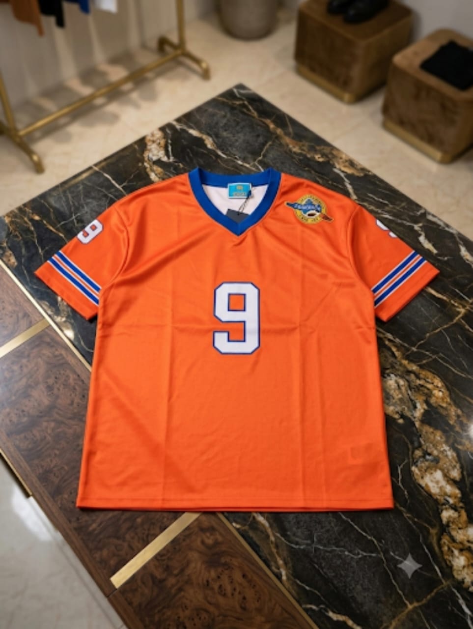 Bright orange V-neck big tee with number 9 and sleeve stripes displayed on marble table.