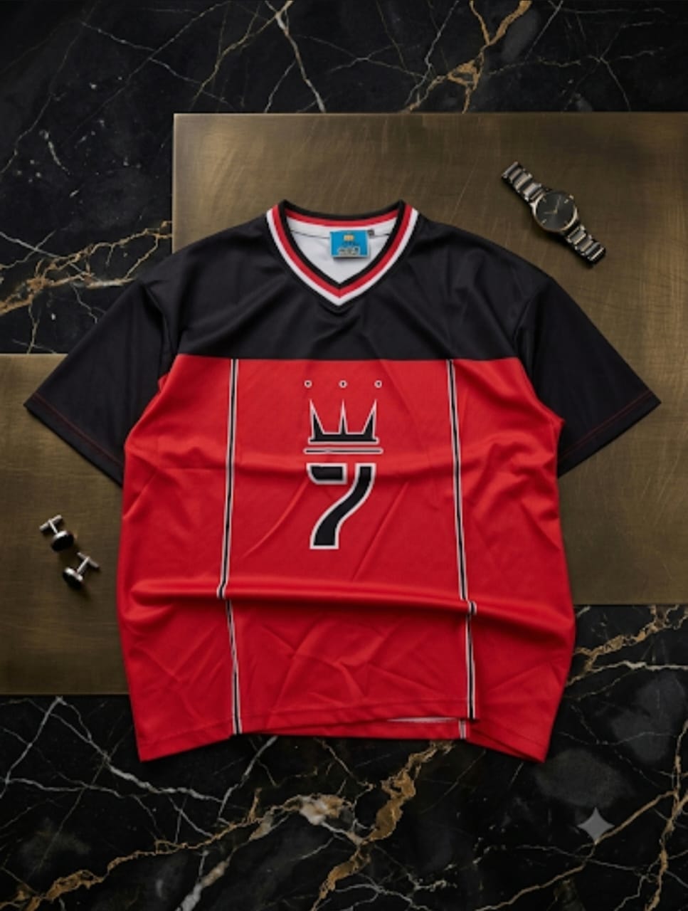 Red and black colorblock V-neck big tee with crown and number 7 graphic laid flat on marble surface with accessories.