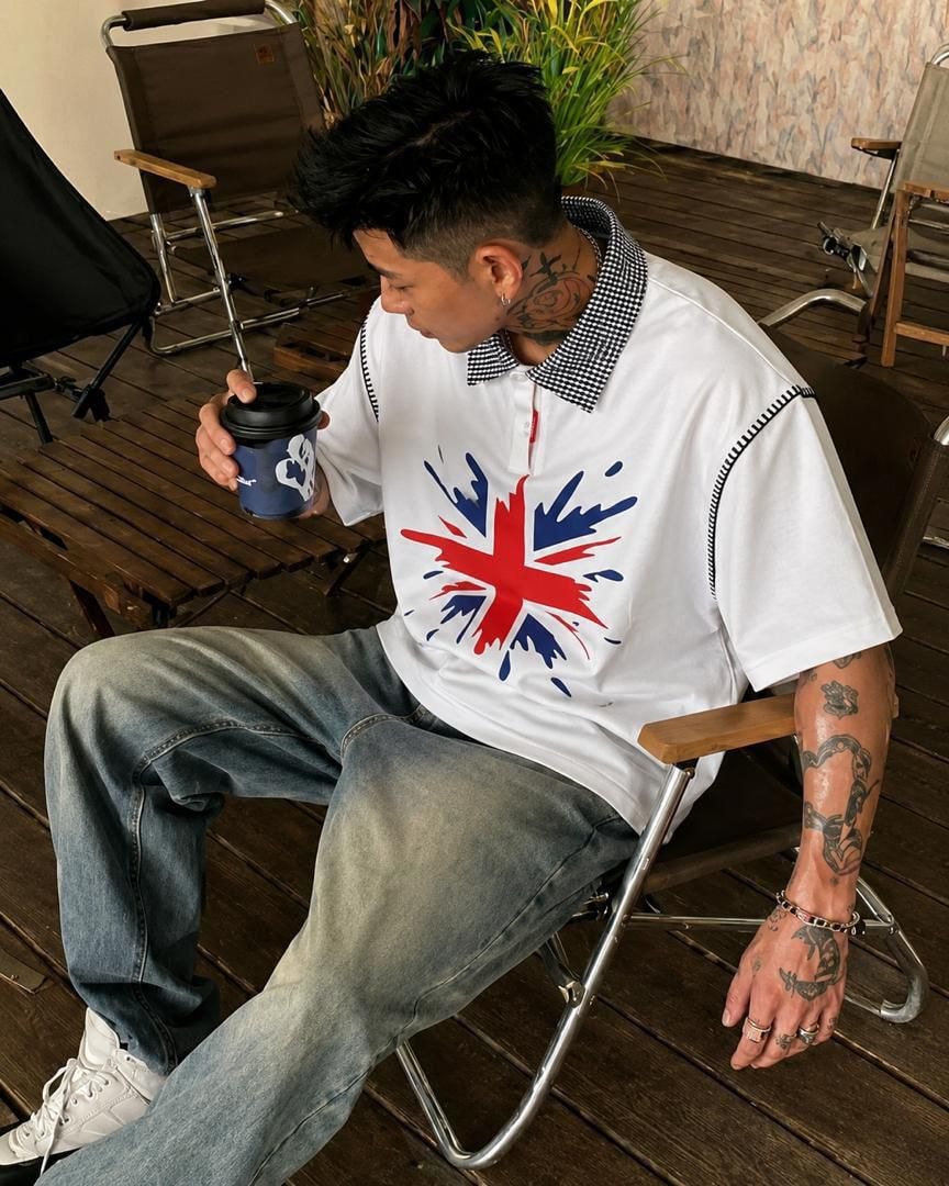 Man with tattoos sitting outdoors wearing white Union Jack splatter polo shirt with checkered collar and jeans.