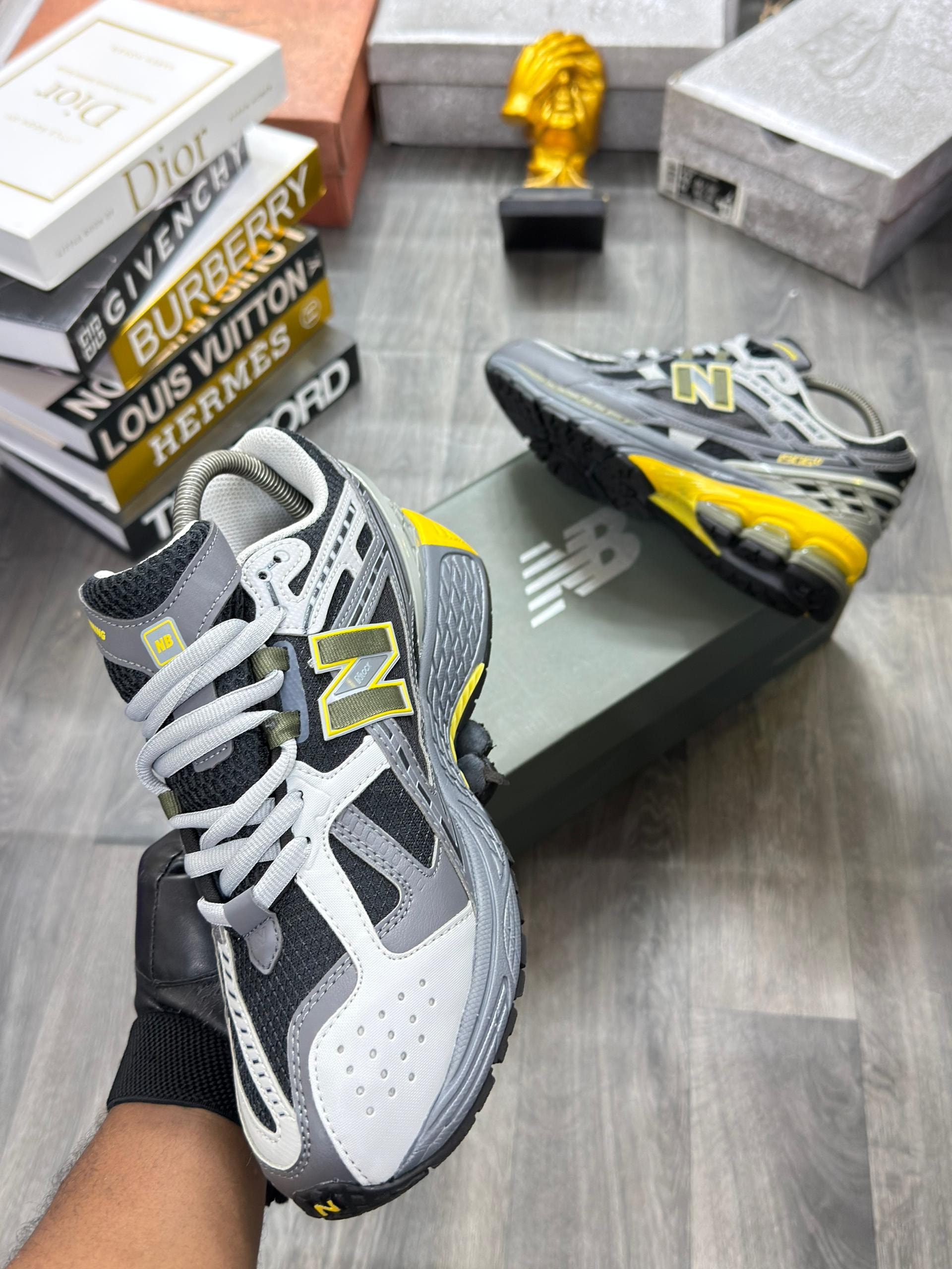Gray yellow and black New Balance sneakers held in gloved hand with box and books behind.