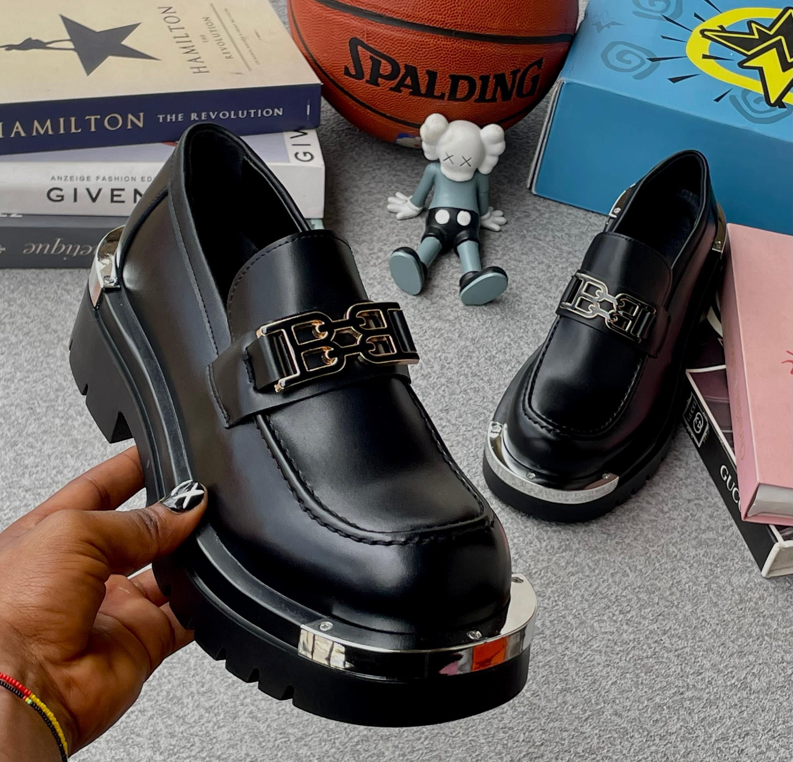 Black platform loafers with large metal buckle held by hand with lifestyle props in background.