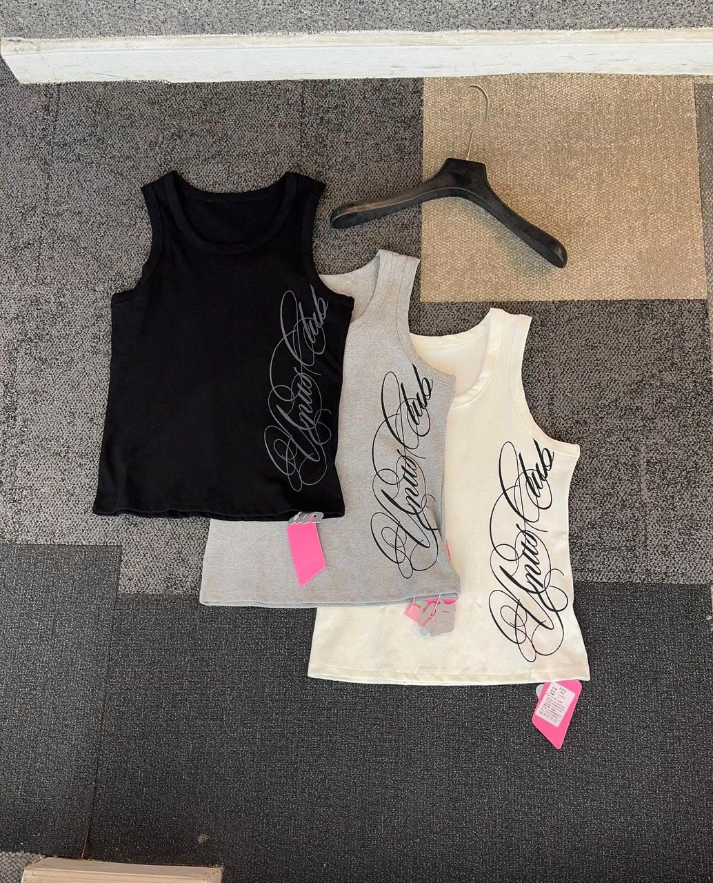 Three armless tank tops in black, grey and white laid out together showing side script prints.