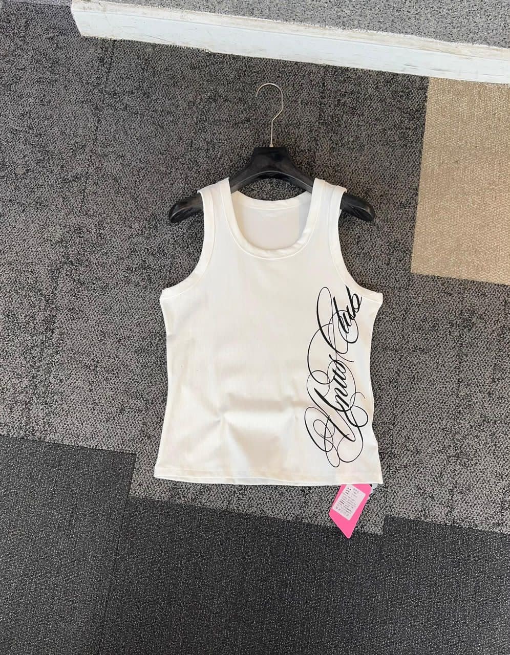 White armless tank top with black cursive script print on the side, hanging on a black hanger.
