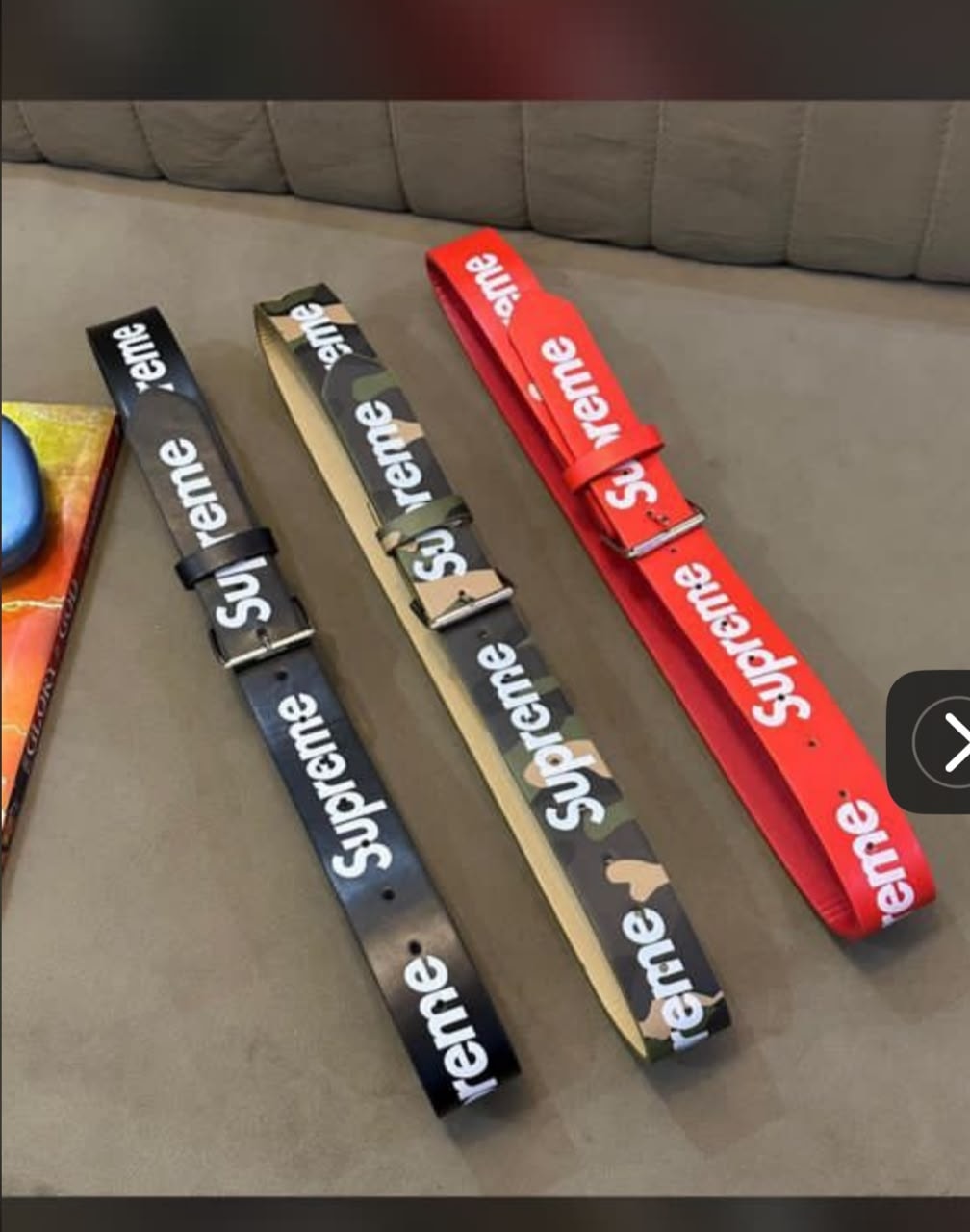 Three Supreme webbing belts in black, camouflage, and red with logo prints and buckles laid on a surface.