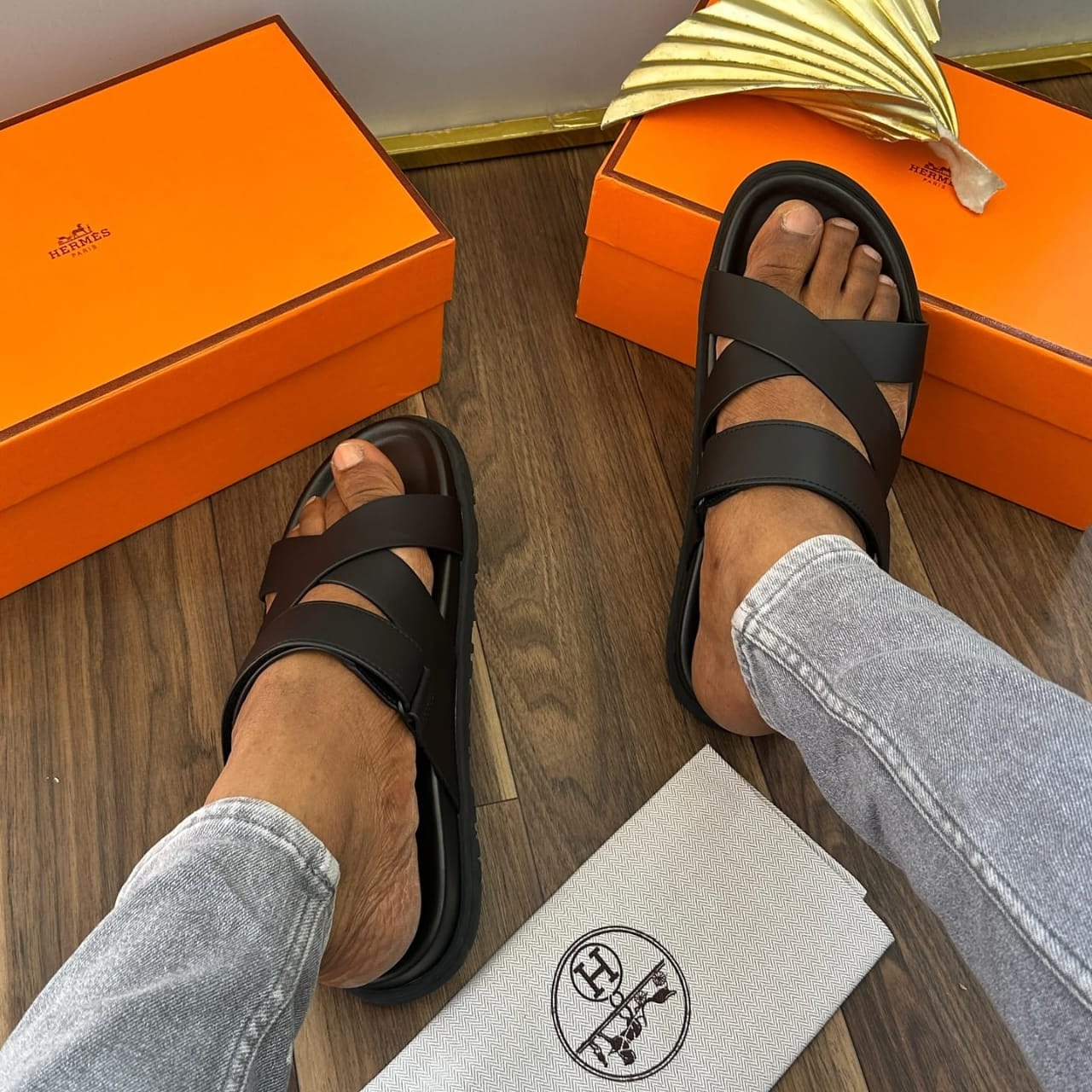 Person wearing black Hermes cross strap sandals with orange boxes and dust bag.