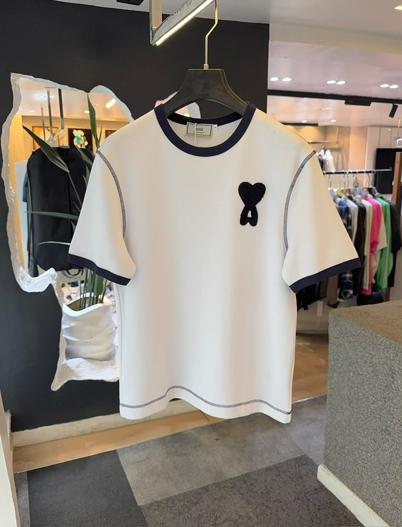 White AMI polo shirt with navy contrast trim and black heart logo hanging in store.