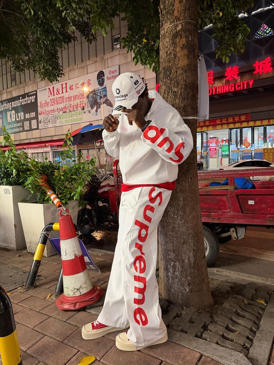 Model in white Supreme two-piece tracksuit leaning against a tree at night.