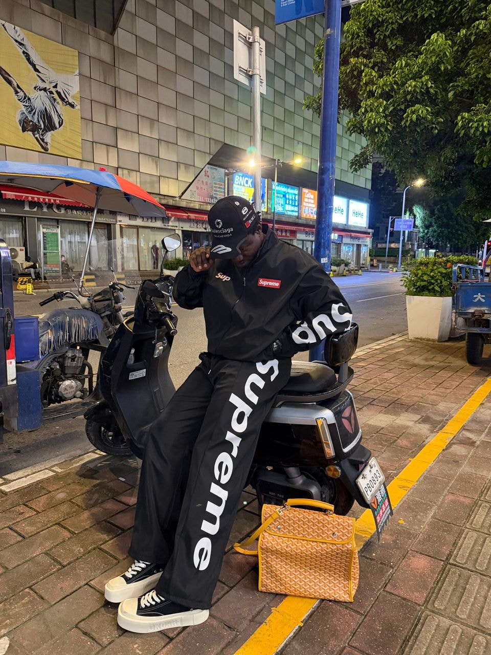 Man in black Supreme tracksuit set sitting on a scooter at night.