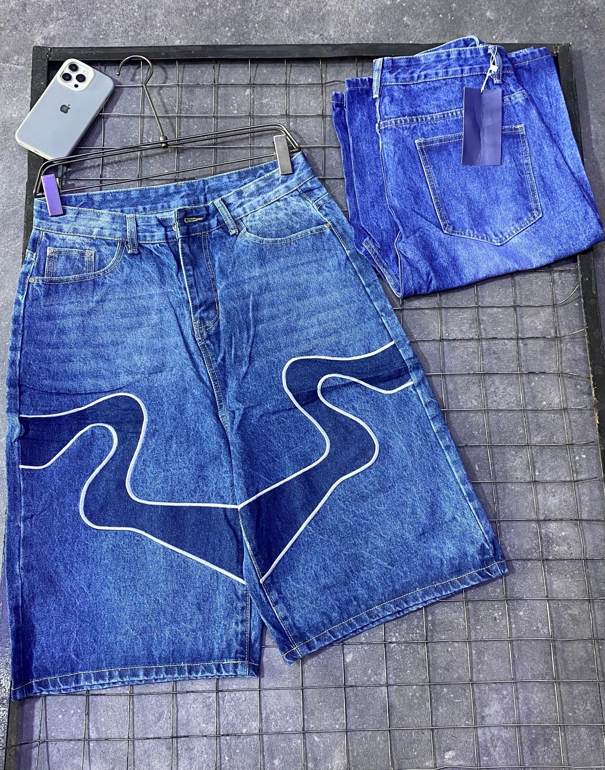 Blue denim jorts with white squiggle wave design on front, displayed on hanger beside folded pair and phone.