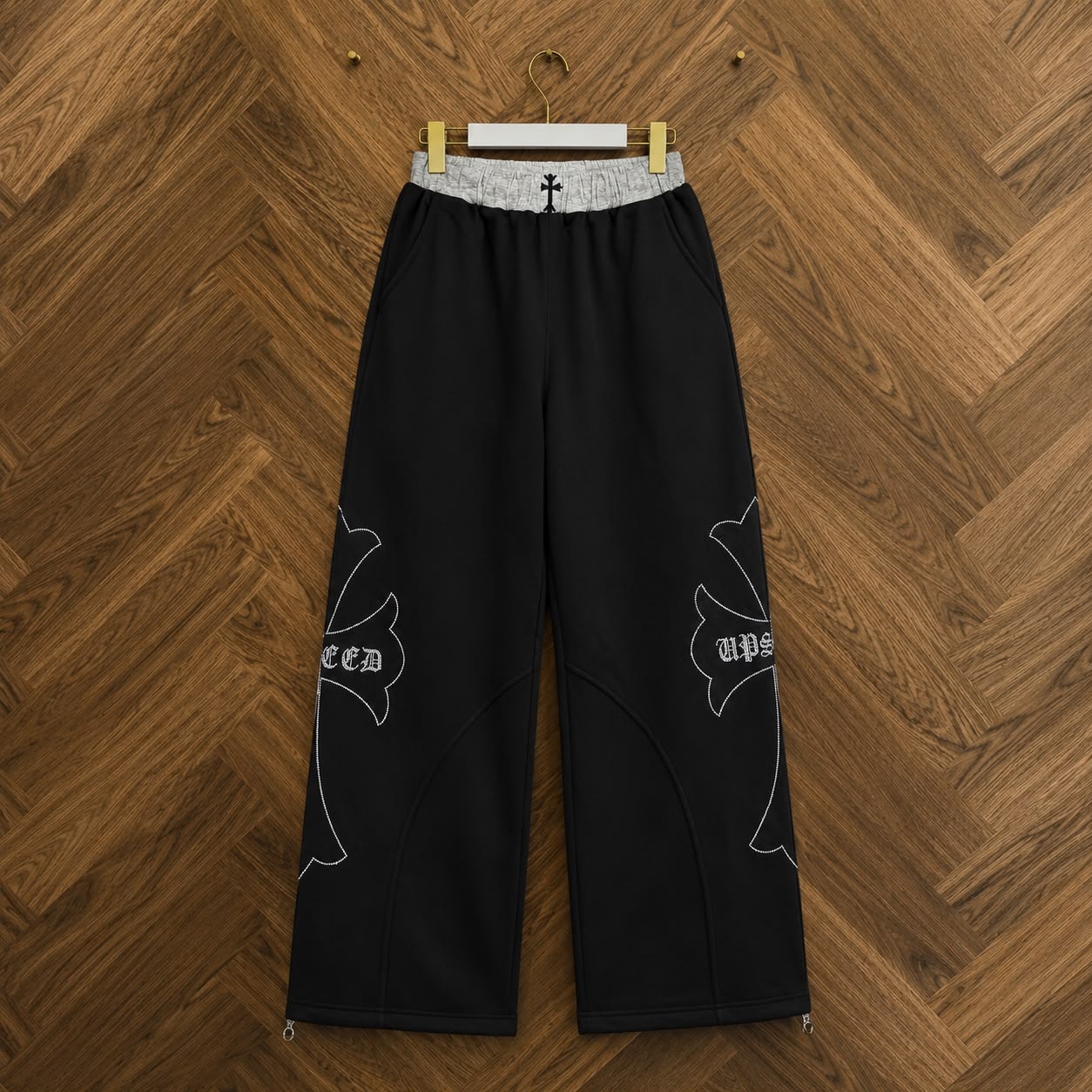 Black joggers with white embroidered designs containing gothic text on the legs and grey waistband with cross, hanging on hanger.