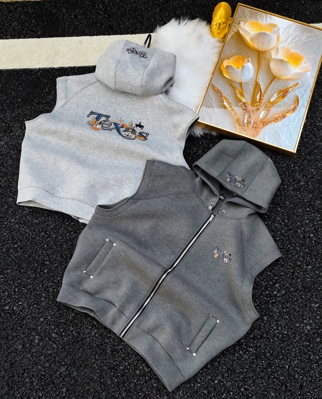 Sleeveless hoodies with Texas embroidery in light and dark gray beside white fur and golden flowers.