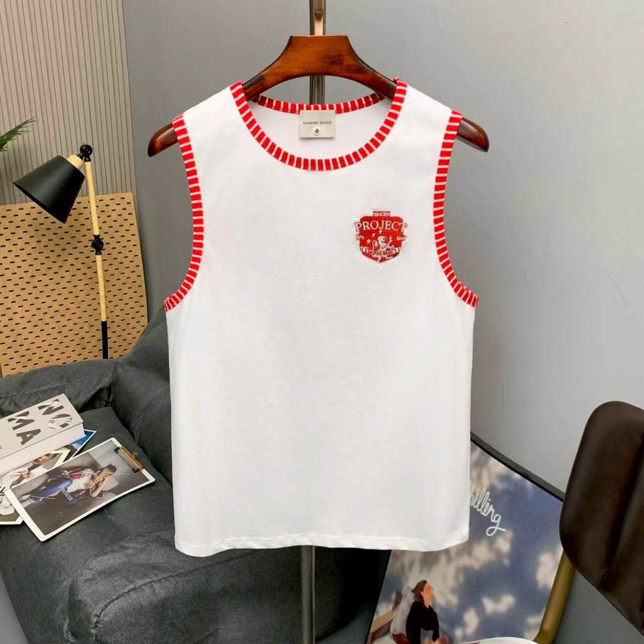 White armless tank top with red striped trim and chest logo on wooden hanger in room setting.