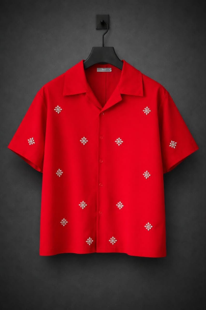 Red short sleeve button-up shirt with scattered silver snowflake embroidery, hanging on black hanger against dark background.