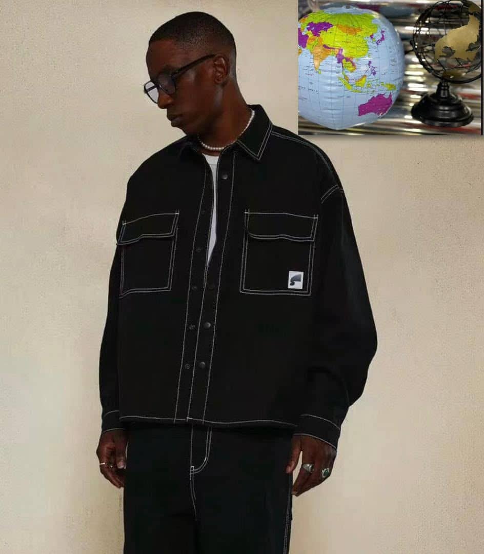 Man wearing black long sleeve contrast stitch shirt with chest pockets, styled with glasses and accessories against neutral wall.