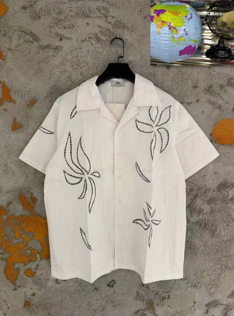 White short sleeve button-up shirt with black dotted floral and leaf patterns, hanging on a black hanger against a textured wall with decorative globe.