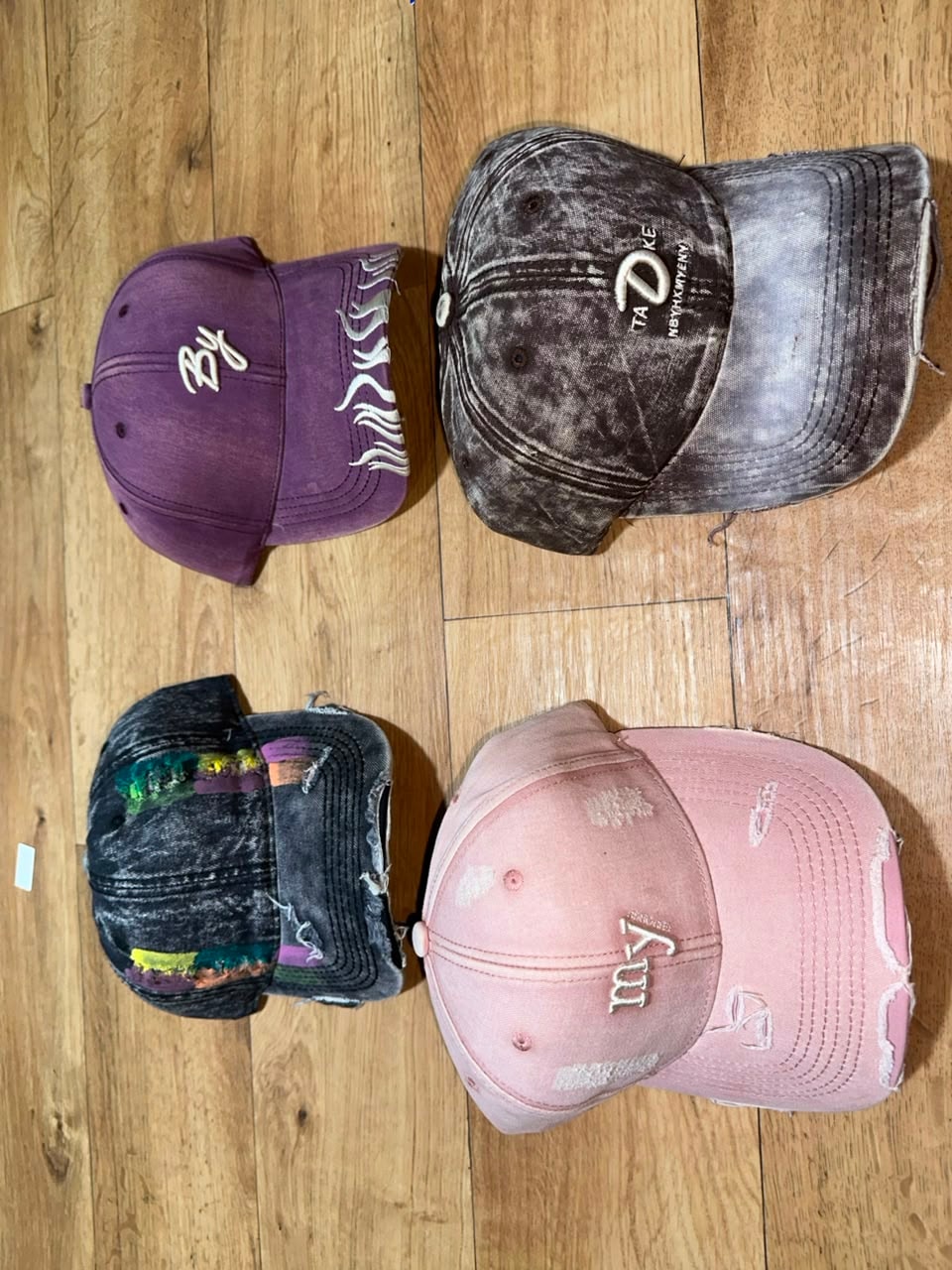 Assorted distressed embroidered baseball caps in purple, gray, black, and pink with various logos and prints.