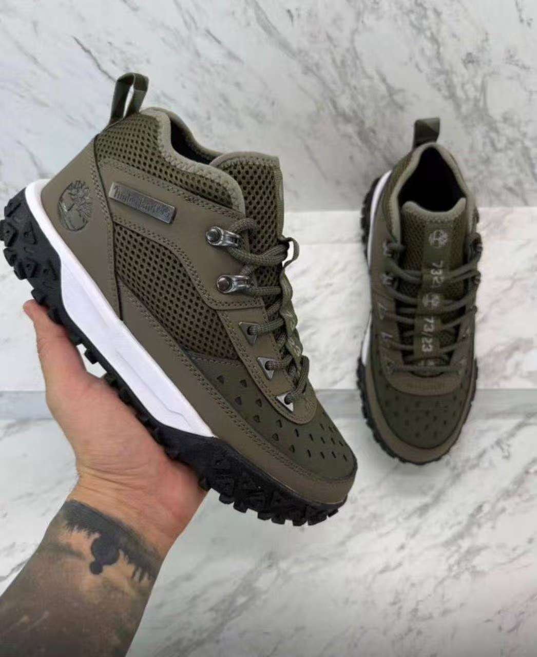Olive green Timberland hiking shoes held in hand and styled on marble background.