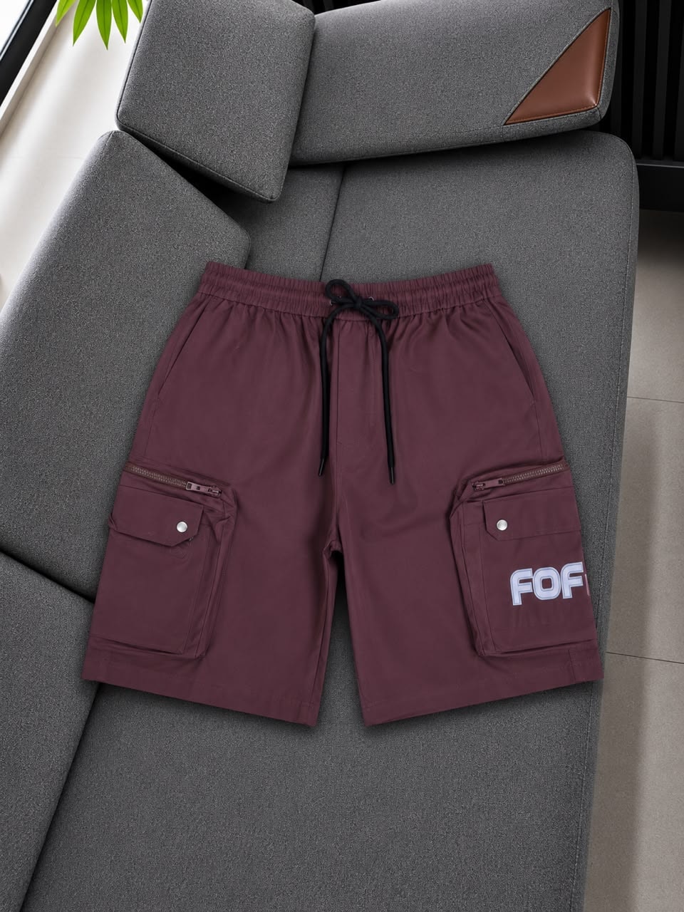 Burgundy FOF cargo shorts displayed on gray sofa with visible logo and pockets.