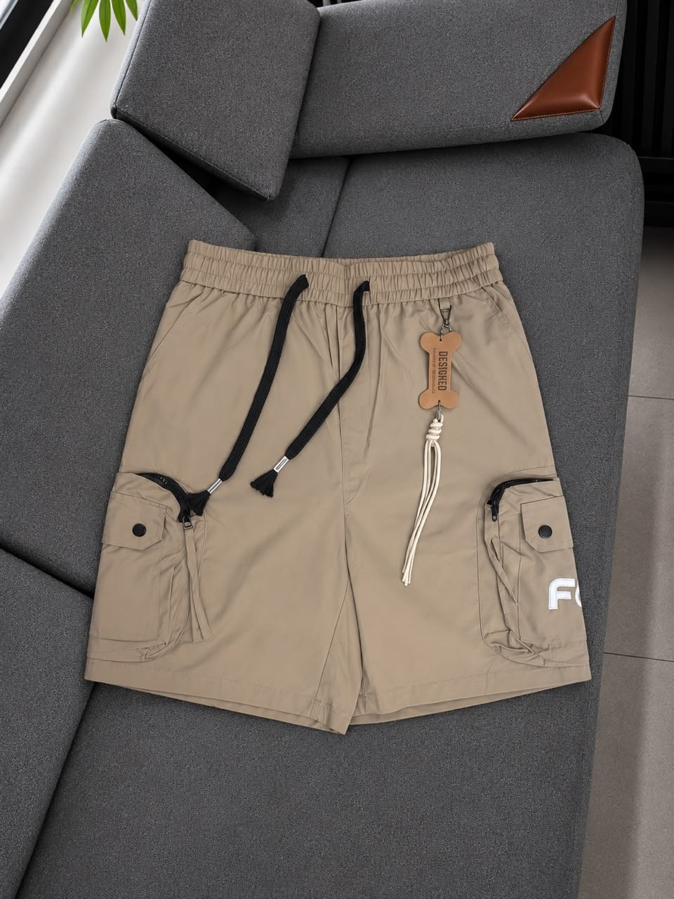 Beige FOF cargo shorts on gray couch with black drawstring and logo detail.