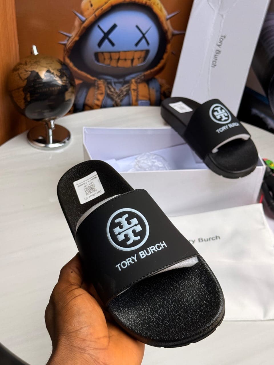 Black Tory Burch slide held in hand displaying logo, sole and packaging on white table.
