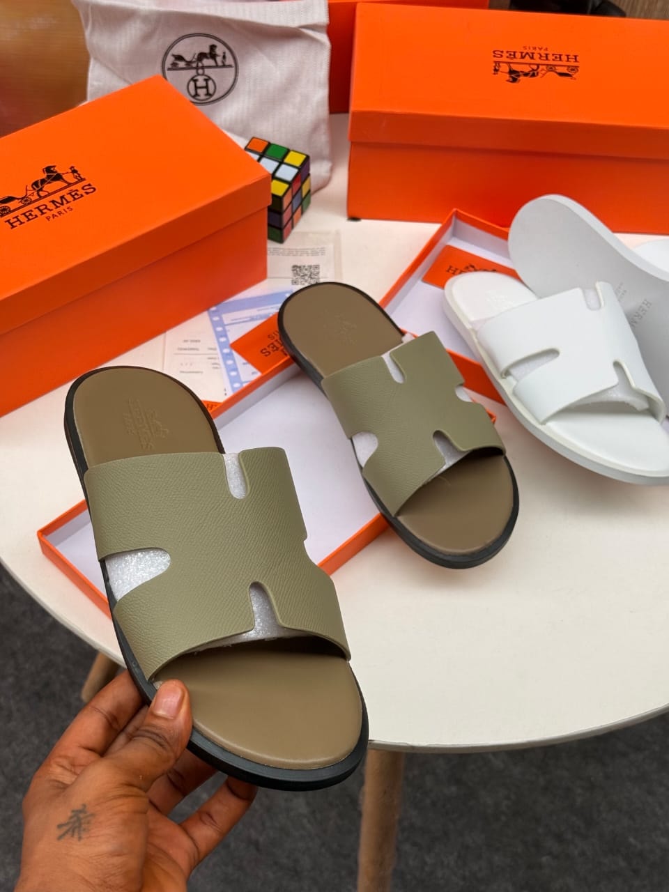 Hermes H-strap slides shown in olive and white colorways with orange boxes and accessories.