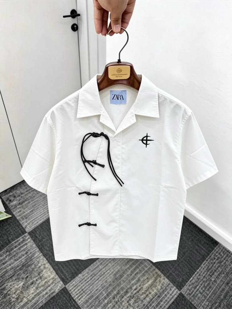White short sleeve shirt with frog buttons, drawstring and chest embroidery on wooden hanger.