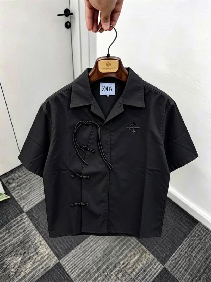 Black short sleeve shirt with frog buttons, drawstring and chest embroidery on wooden hanger.