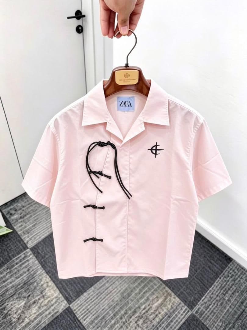 Pink short sleeve shirt with frog buttons, drawstring and chest embroidery on wooden hanger.