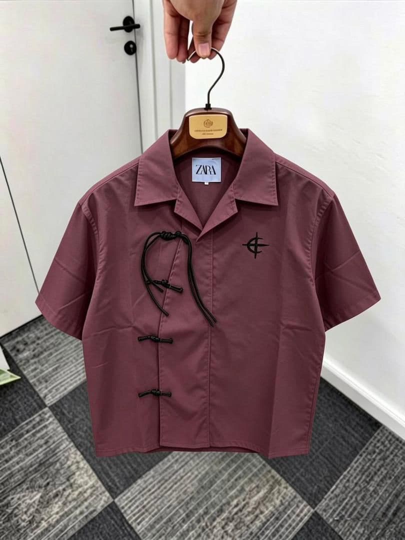 Burgundy short sleeve shirt with frog buttons, drawstring and chest embroidery on wooden hanger.