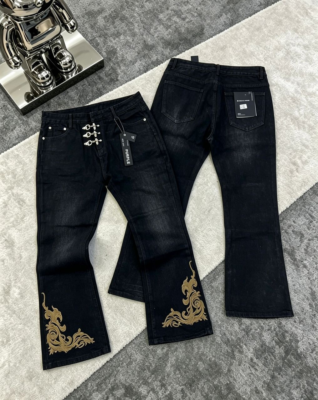 Black flare jeans with orange swirl embroidery on the thigh, hanging on a rack with tags.
