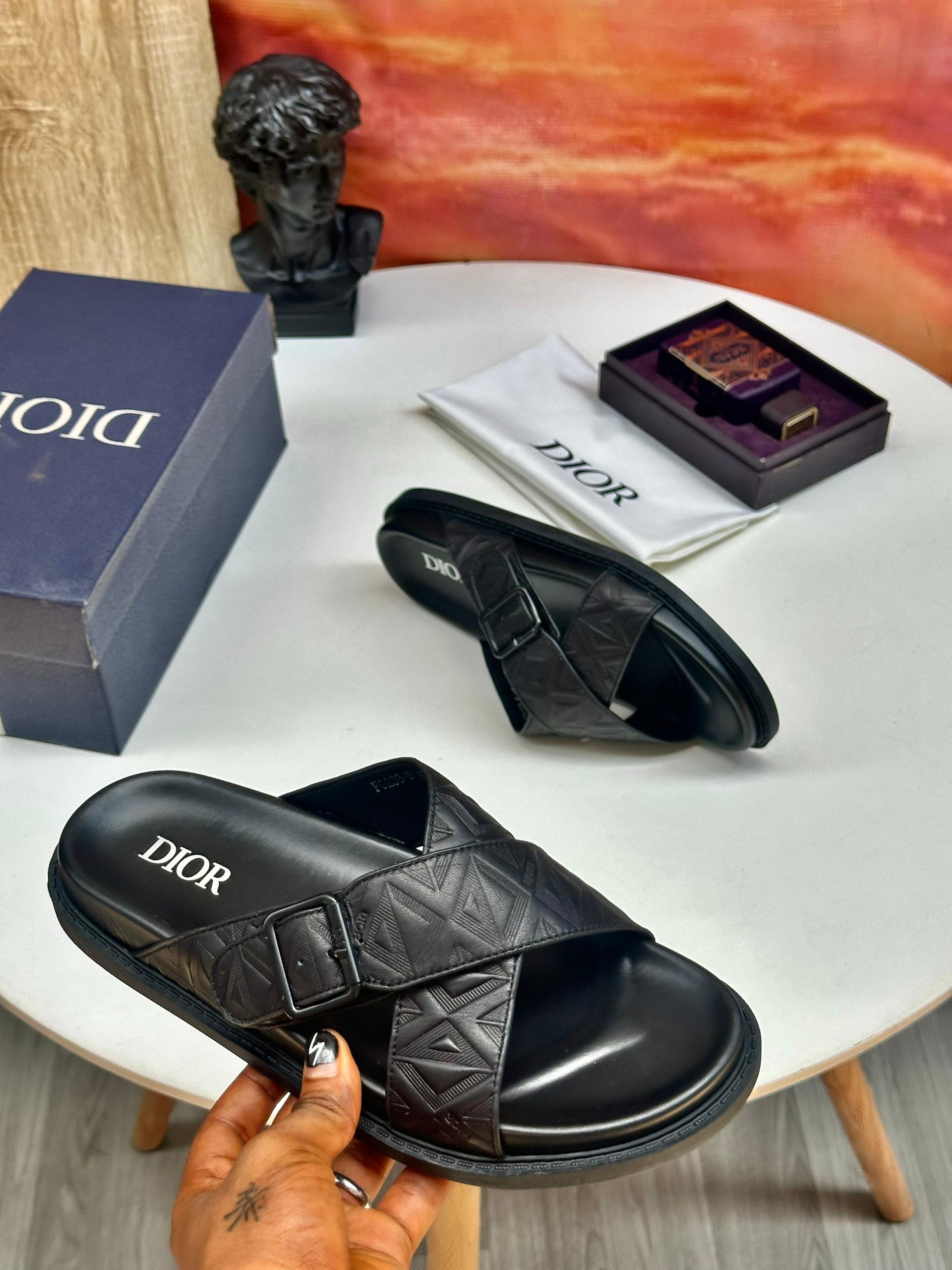 Black Dior slides with patterned strap and buckle shown with Dior box, dust bag and perfume on white table.