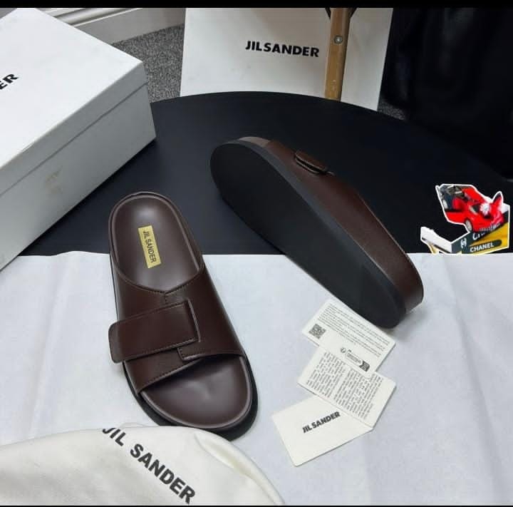 Brown Jil Sander single strap slide with buckle on white surface with brand packaging and cards.